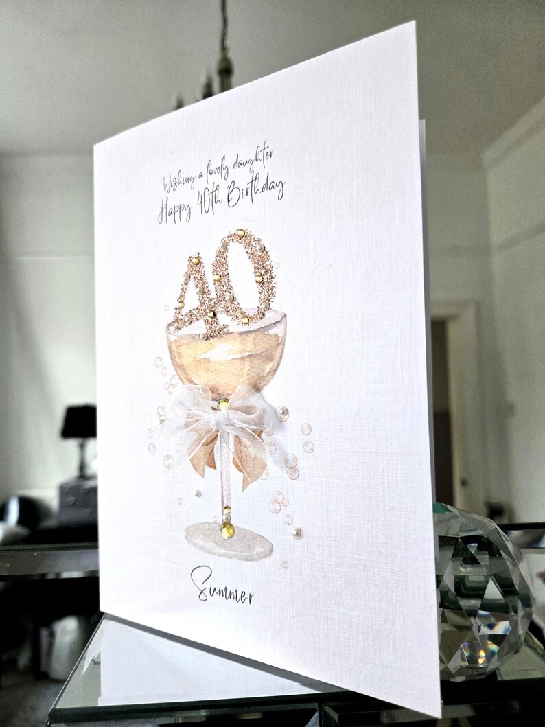 May include: A white birthday card with the text "Wishing a lovely daughter Happy 40th Birthday." The card features a champagne glass with a "40" made of gold glitter. A white bow and pearls decorate the glass. The name "Summer" is written at the bottom.