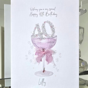 May include: A white greeting card with the text "Wishing you a very special Happy 40th Birthday." The card features a champagne glass design with a pink bow and the name "Lilly" at the bottom. The number "40" is decorated with silver glitter.