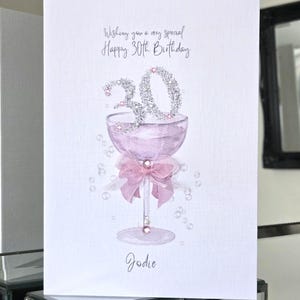 Op de afbeelding: Een witte wenskaart met een illustratie van een lichtpaars cocktailglas met een roze strik, het getal 30 in zilveren glitters en kleine doorzichtige bubbels. De tekst luidt: Wishing you a very special Happy 30th Birthday en de naam Jodie.
