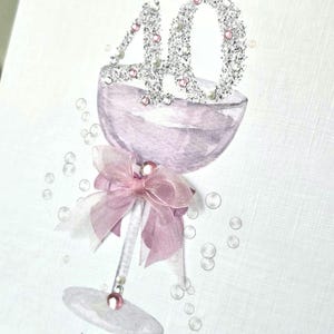 May include: A greeting card with a champagne glass design. The number "40" is embellished with glitter and pearls. A pink bow adorns the glass stem. The words "Happy 40th Birthday" and the name "Lilly" are written on the card.