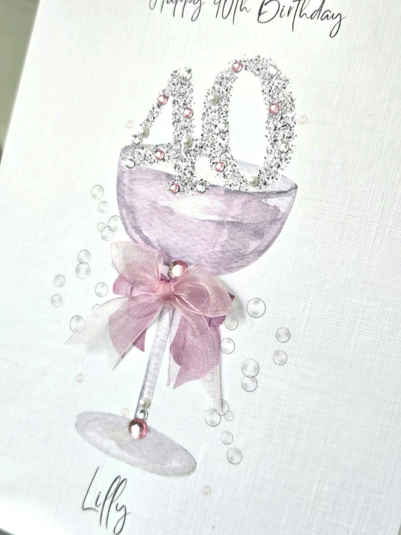 May include: A greeting card with a champagne glass design. The number "40" is embellished with glitter and pearls. A pink bow adorns the glass stem. The words "Happy 40th Birthday" and the name "Lilly" are written on the card.