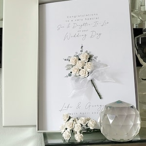 Puede incluir: Tarjeta blanca con un ramo de flores blancas y el texto "Felicidades a un hijo y una nuera muy especiales en su d&iacute;a de boda" y "Luke & Georgie 9 de abril de 202".