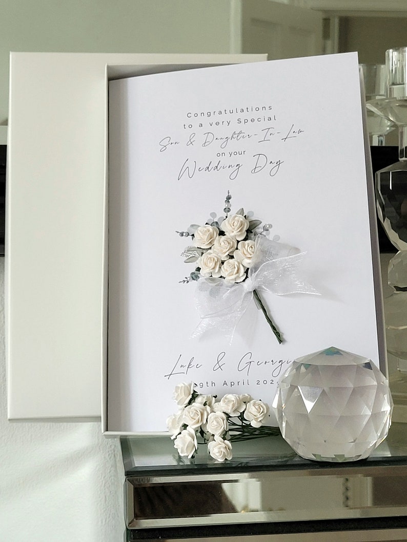 Puede incluir: Tarjeta blanca con un ramo de flores blancas y el texto "Felicidades a un hijo y una nuera muy especiales en su d&iacute;a de boda" y "Luke & Georgie 9 de abril de 202".