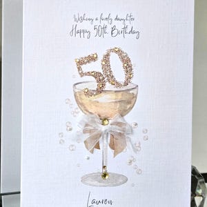 Op de afbeelding: Een witte wenskaart met de tekst "Wishing a lovely daughter Happy 50th Birthday". De kaart toont een champagneglas met een glitterende "50" erop. Een strik en parels versieren het glas. De naam "Lauren" staat onderaan.