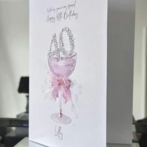 May include: A white birthday card with a watercolor illustration of a champagne glass filled with pink liquid. The number "40" is at the top, and a pink bow adorns the base. The card reads "Wishing you a very special Happy 40th Birthday."