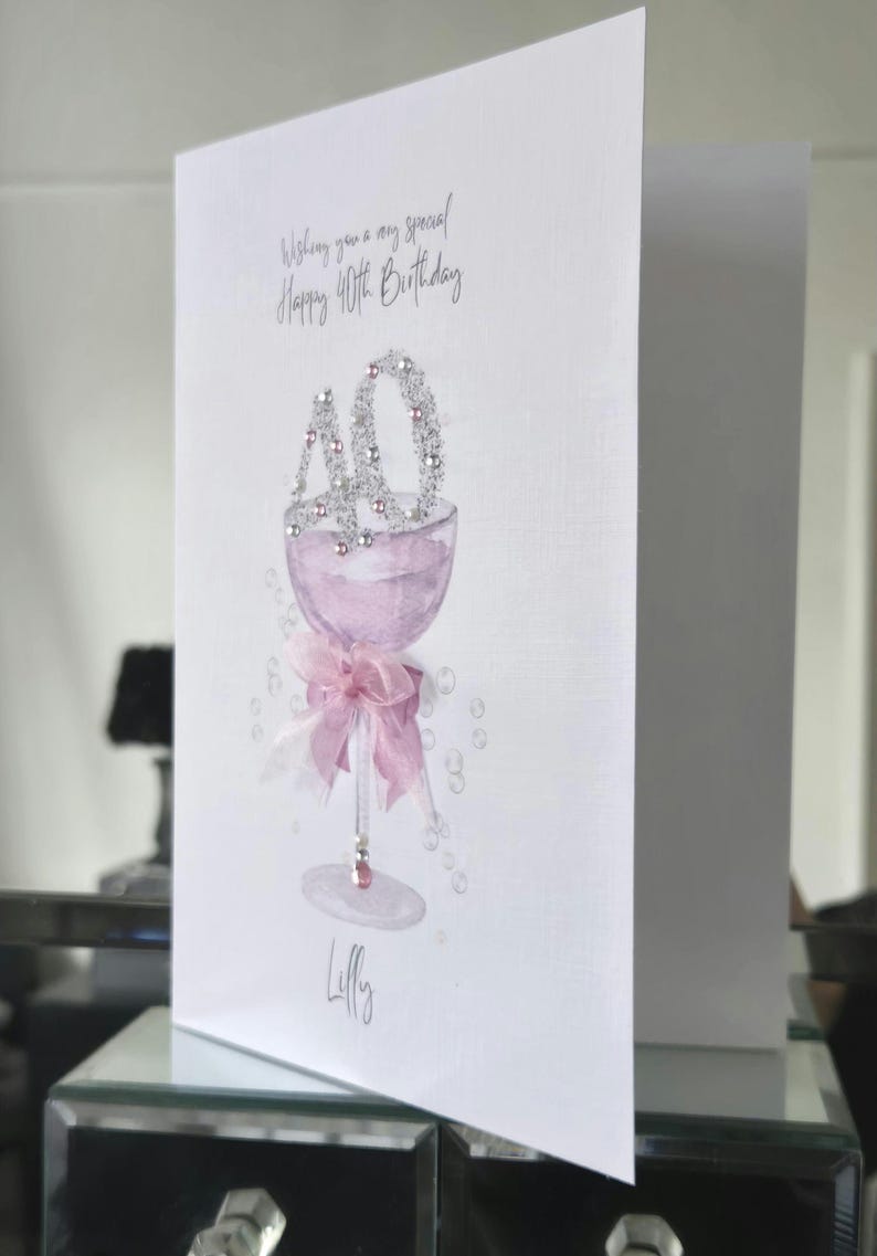 May include: A white birthday card with a watercolor illustration of a champagne glass filled with pink liquid. The number "40" is at the top, and a pink bow adorns the base. The card reads "Wishing you a very special Happy 40th Birthday."