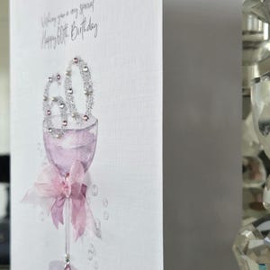 May include: A white greeting card with a watercolor illustration of a wine glass. The glass is filled with a light purple liquid and adorned with a pink bow. The number "60" is embellished with silver glitter. The text reads "Wishing you a very special Happy 60th Birthday."