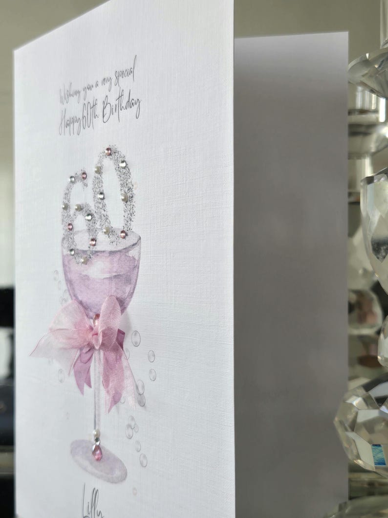 May include: A white greeting card with a watercolor illustration of a wine glass. The glass is filled with a light purple liquid and adorned with a pink bow. The number "60" is embellished with silver glitter. The text reads "Wishing you a very special Happy 60th Birthday."