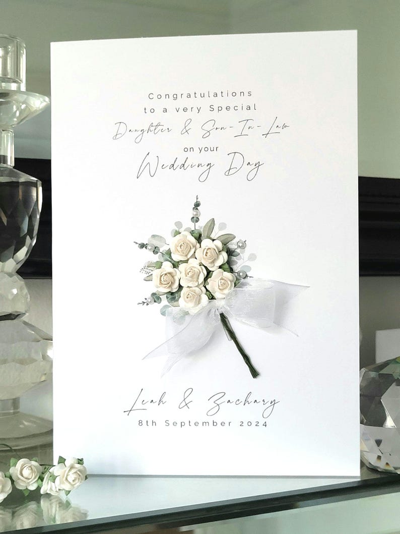 Puede incluir: Tarjeta de boda blanca con un ramo de flores blancas y hojas verdes. La tarjeta dice "Felicidades a una hija y un yerno muy especiales por su d&iacute;a de boda" y "Leah & Zachary 8 de septiembre de 2024".