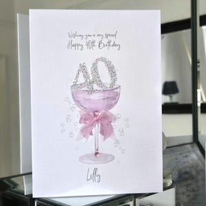 May include: A greeting card with the text "Wishing you a very special Happy 40th Birthday." The card features a watercolor illustration of a champagne glass with the number "40" in rhinestones. A pink bow adorns the glass stem, and the name "Lilly" is written below.