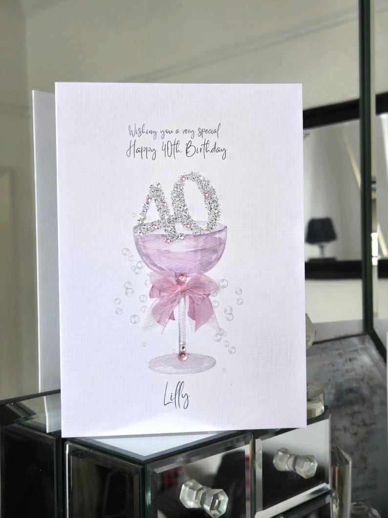 May include: A greeting card with the text "Wishing you a very special Happy 40th Birthday." The card features a watercolor illustration of a champagne glass with the number "40" in rhinestones. A pink bow adorns the glass stem, and the name "Lilly" is written below.
