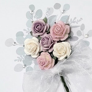 Peut inclure: Un bouquet de fleurs en papier dans des tons de rose, blanc et violet, nou&eacute; d'un ruban blanc transparent. Les fleurs sont dispos&eacute;es en grappe et entour&eacute;es de feuilles vertes.