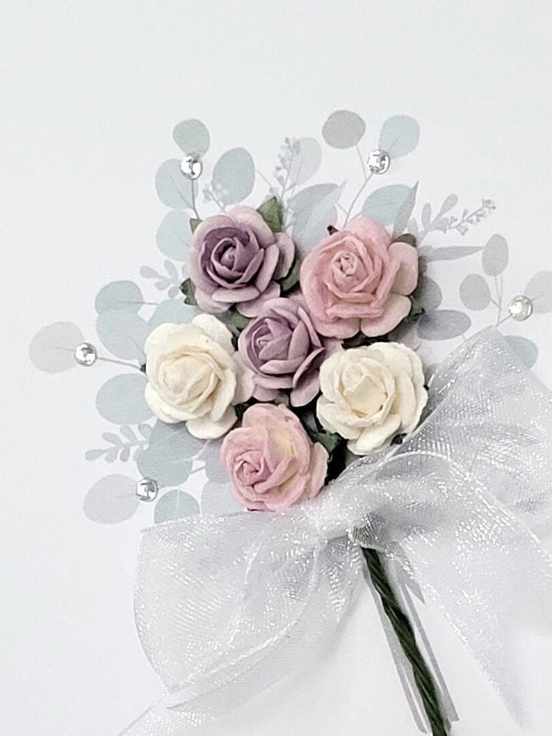 Peut inclure: Un bouquet de fleurs en papier dans des tons de rose, blanc et violet, nou&eacute; d'un ruban blanc transparent. Les fleurs sont dispos&eacute;es en grappe et entour&eacute;es de feuilles vertes.