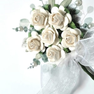 Puede incluir: Un peque&ntilde;o ramo de rosas de papel blanco con hojas verdes y un lazo de tul blanco. El ramo est&aacute; atado con una cinta blanca.