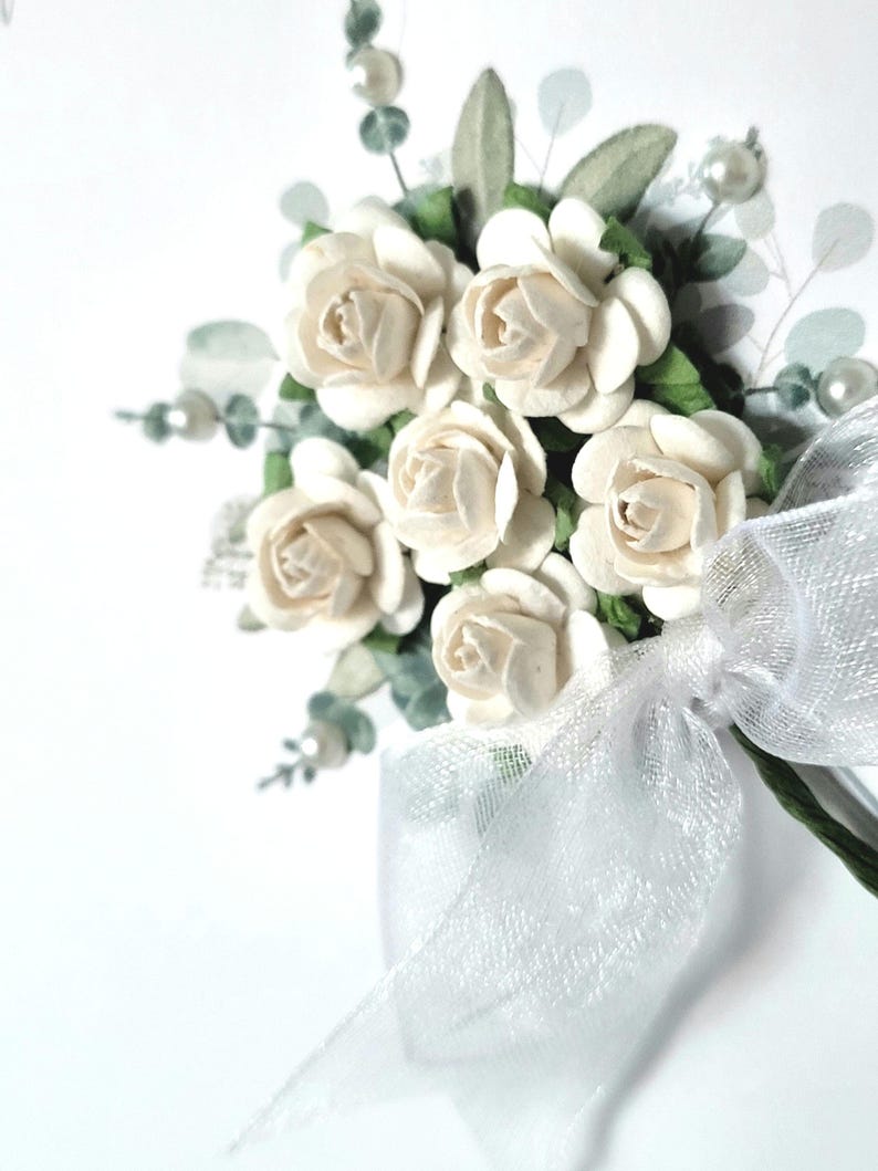 Puede incluir: Un peque&ntilde;o ramo de rosas de papel blanco con hojas verdes y un lazo de tul blanco. El ramo est&aacute; atado con una cinta blanca.