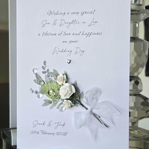 Puede incluir: Una tarjeta de boda blanca con el texto "Wishing a very special Son & Daughter in law a lifetime of love and happiness on your Wedding Day". Presenta un arreglo floral con rosas blancas y una cinta blanca. Los nombres "Sarah & Jack" y la fecha "24 de febrero de 2023" también están impresos.
