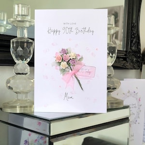 May include: A white card with pink and white flowers and a pink ribbon. The card says "Happy 90th Birthday" and "With Love" and "Mum".