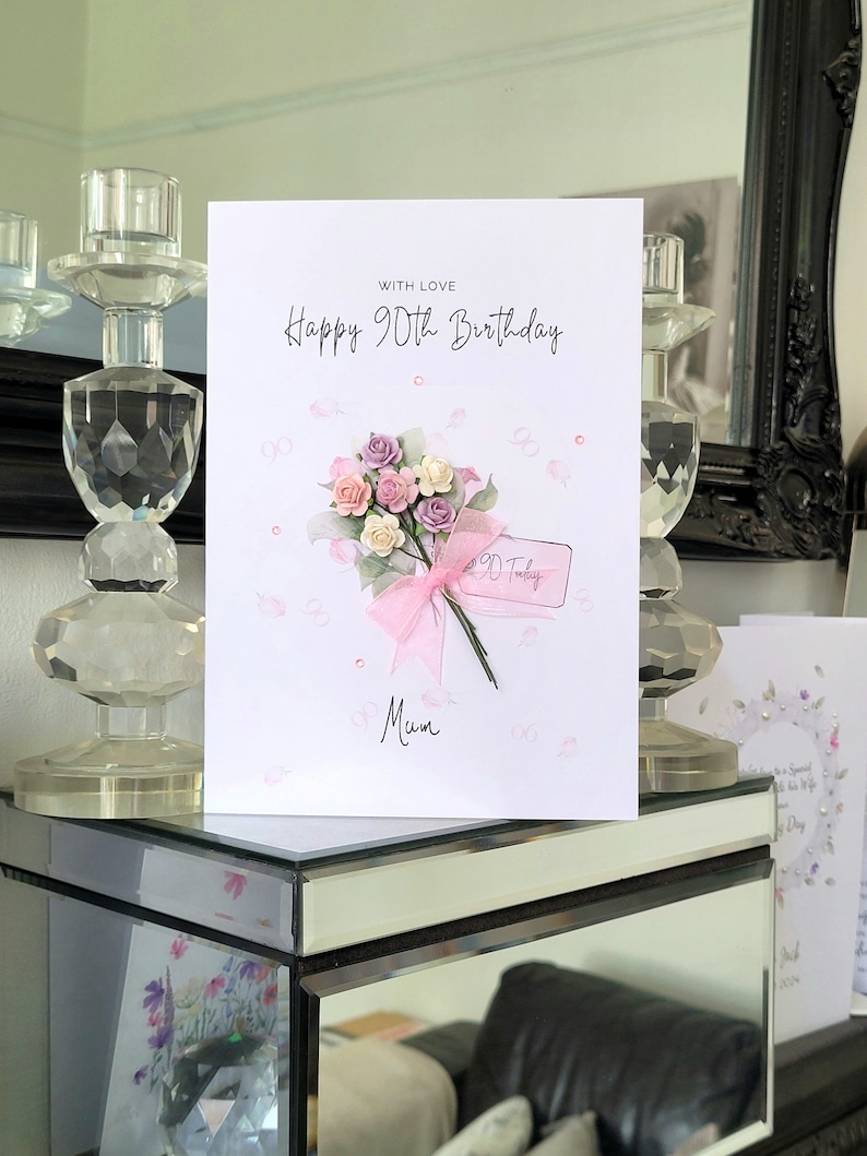 May include: A white card with pink and white flowers and a pink ribbon. The card says "Happy 90th Birthday" and "With Love" and "Mum".