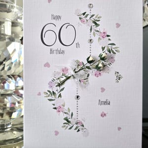 Op de afbeelding: Een witte 60e verjaardagskaart met de tekst "Happy 60th Birthday" in zwarte letters. De kaart is versierd met roze rozen, groene bladeren en een rij strass-steentjes. De naam "Amelia" is in script geschreven.