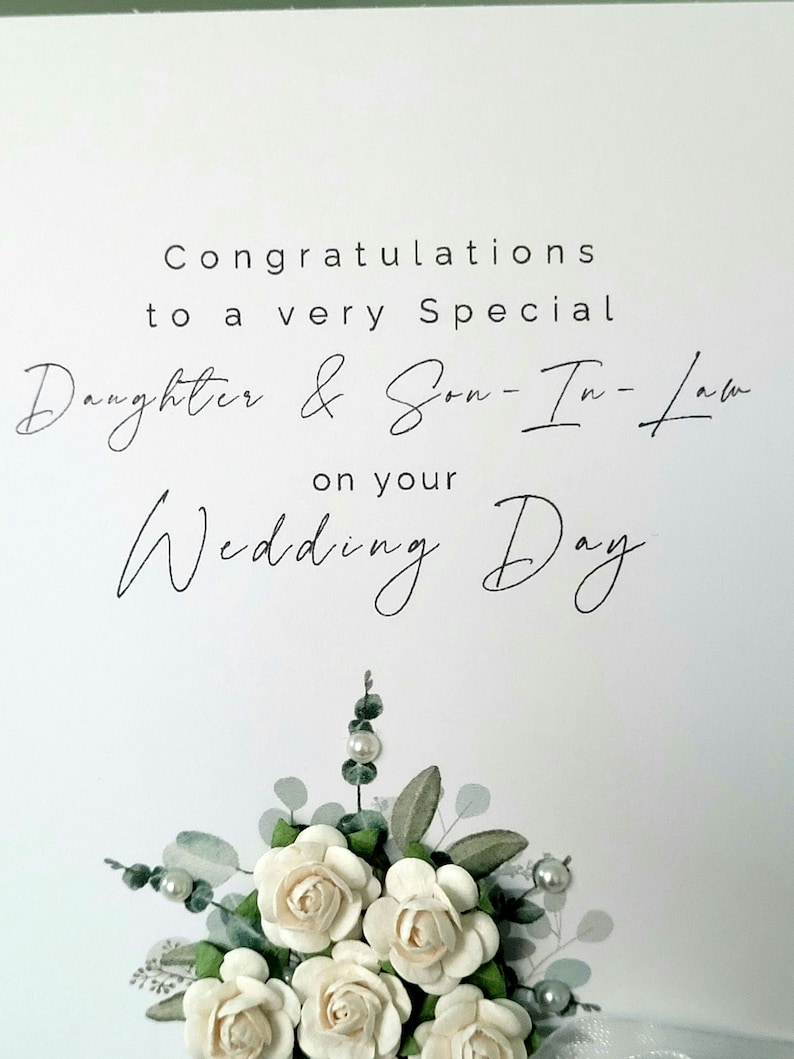 Puede incluir: Una tarjeta blanca con caligraf&iacute;a negra que dice "Felicidades a una hija y un yerno muy especiales por su d&iacute;a de boda". La tarjeta est&aacute; decorada con un ramo de rosas de papel blanco y hojas verdes.