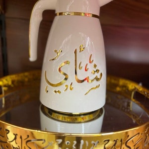 May include: White thermal carafe with a gold lid and trim. The carafe has Arabic calligraphy in gold on the side that reads "Shai". The carafe is sitting on a gold tray with Arabic calligraphy.
