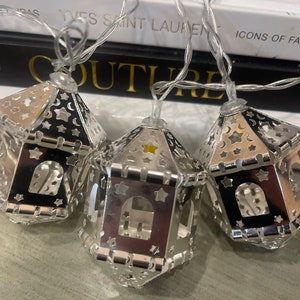 May include: Silver string lights with decorative metal lanterns shaped like stars and crescent moons. The lanterns have a cut-out design and are illuminated with warm white LED lights.