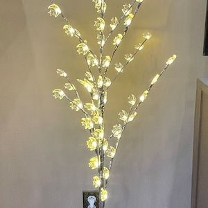 May include: A silver metal floor lamp with a tree-like design featuring crescent moon-shaped lights. The lamp is lit and has a white cord. Two gold lanterns with red candles are on the floor next to the lamp.