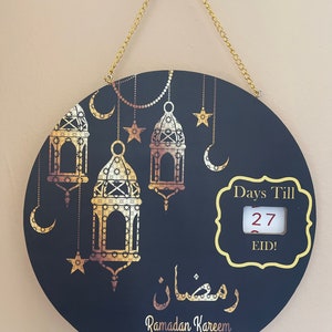 May include: A black circular countdown sign with gold accents. The sign features a crescent moon, stars, and lanterns. The text "Days Till Eid!" is displayed with a number window showing "27". The sign also includes the Arabic text "رمضان" and the phrase "Ramadan Kareem".