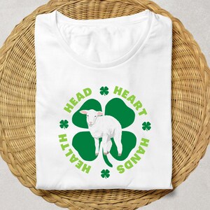 May include: White t-shirt with a green four-leaf clover design. The clover has a white lamb inside and the words "Head, Heart, Health, Hands" around it.