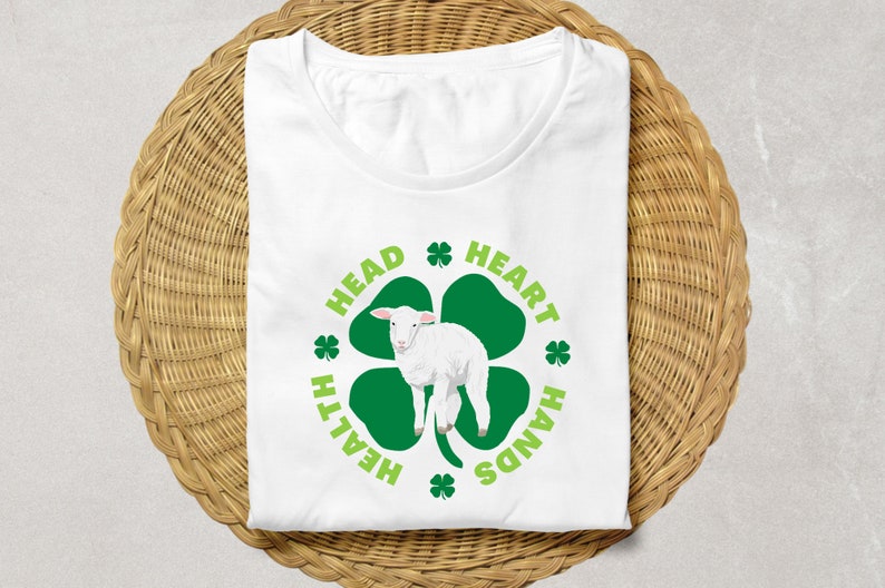 May include: White t-shirt with a green four-leaf clover design. The clover has a white lamb inside and the words "Head, Heart, Health, Hands" around it.