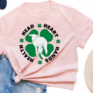 May include: A pink t-shirt with a black and green shamrock design. The shamrock has a white lamb inside and the words "Health", "Head", "Heart", and "Hands" around it.