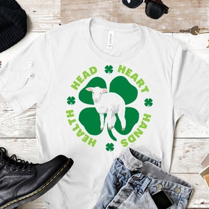 May include: White t-shirt with a green four-leaf clover design. The clover has a white lamb inside and the words "Head, Heart, Hands, Health" around it.