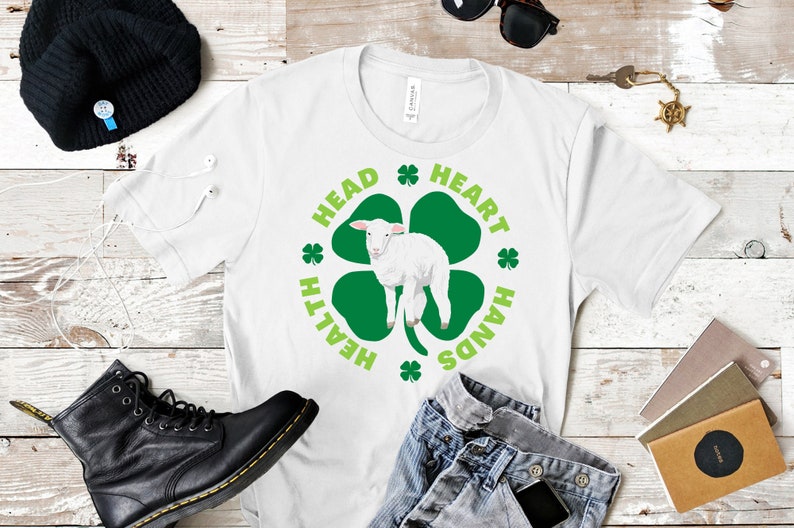 May include: White t-shirt with a green four-leaf clover design. The clover has a white lamb inside and the words "Head, Heart, Hands, Health" around it.