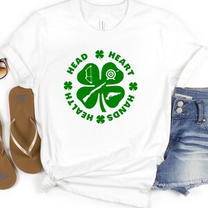 May include: White t-shirt with a green four-leaf clover design. The clover has a bow and arrow, a target, a rifle, and a pistol. The text around the clover reads "Health, Head, Heart, Hands".