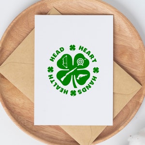May include: A white card with a green four-leaf clover design. The clover has a bow and arrow, a target, a rifle, and a pistol inside. The words "Head, Heart, Hands, Health" are written around the clover.