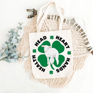 May include: White canvas tote bag with a green four-leaf clover design. The clover has a white lamb in the center. The text around the clover reads "Head, Heart, Hands, Health".