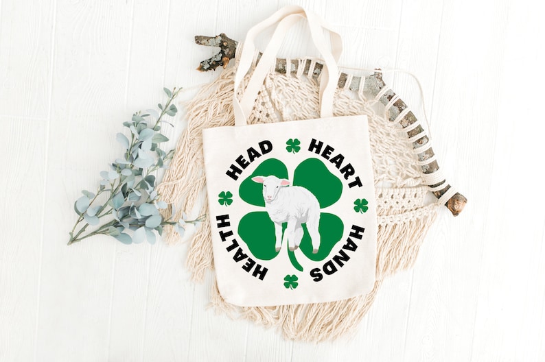 May include: White canvas tote bag with a green four-leaf clover design. The clover has a white lamb in the center. The text around the clover reads "Head, Heart, Hands, Health".