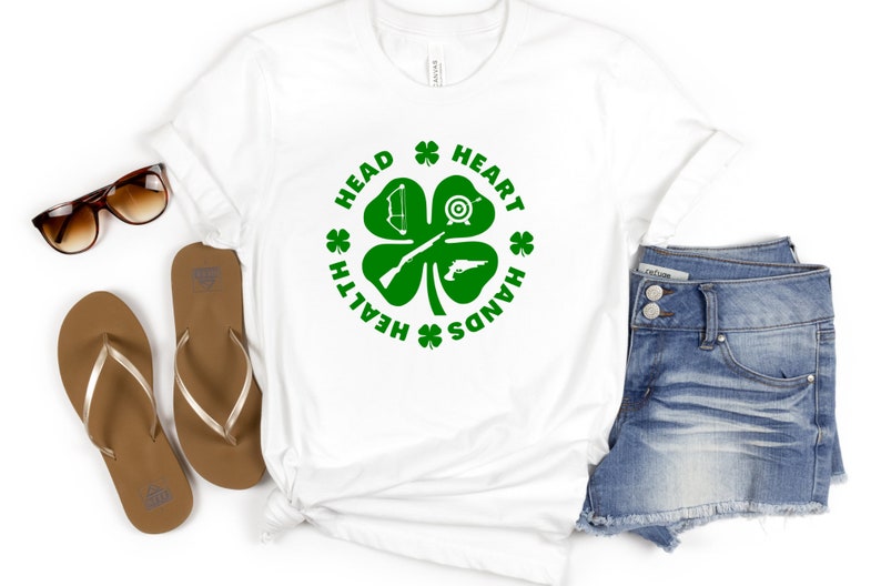 May include: White t-shirt with a green four-leaf clover design. The clover has a bow and arrow, a target, a rifle, and a pistol. The text around the clover reads "Health, Head, Heart, Hands".