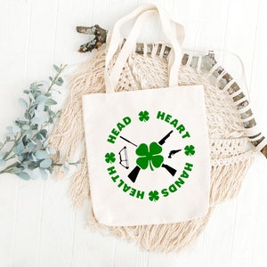 May include: A white canvas tote bag with a green shamrock and the words "Head, Heart, Hands, Health" printed in a circle around it.