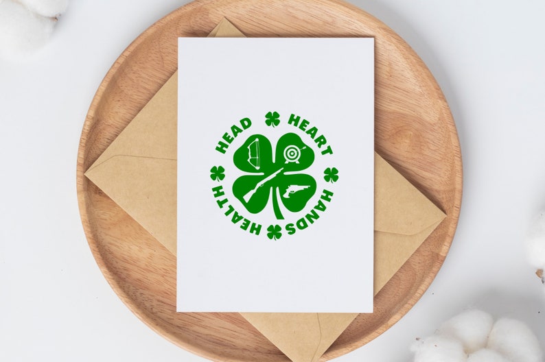 May include: A white card with a green four-leaf clover design. The clover has a bow and arrow, a target, a rifle, and a pistol inside. The words "Head, Heart, Hands, Health" are written around the clover.