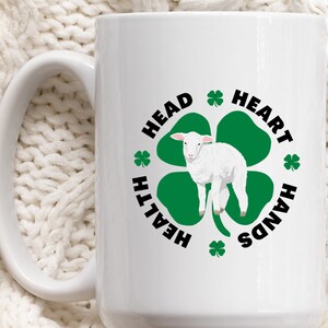 May include: White ceramic mug with a black and green design. The design features a four-leaf clover with a white lamb in the center. The text around the clover reads "Head, Heart, Hands, Health".