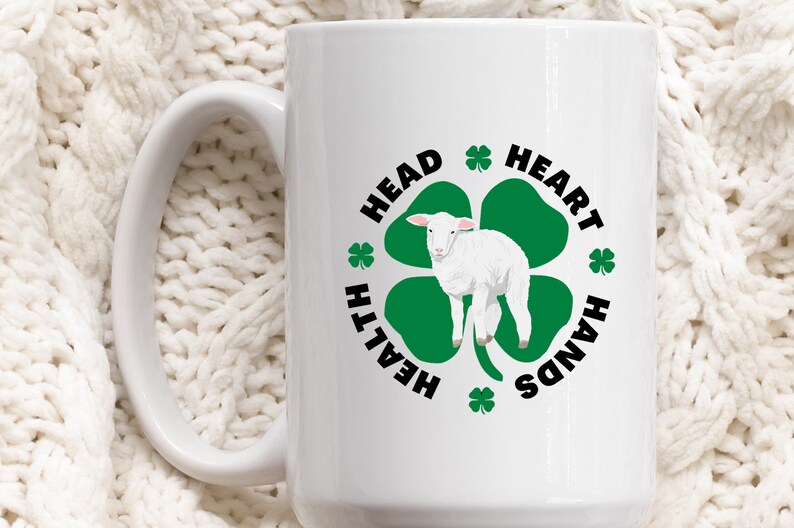 May include: White ceramic mug with a black and green design. The design features a four-leaf clover with a white lamb in the center. The text around the clover reads "Head, Heart, Hands, Health".