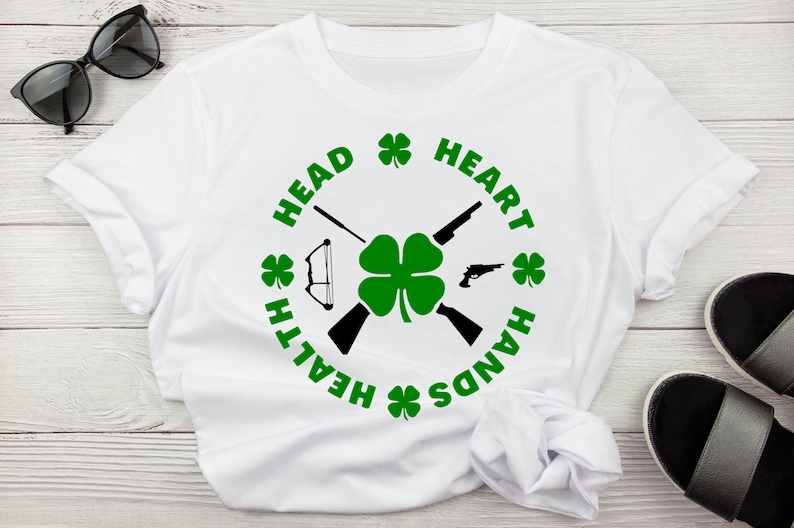 May include: White t-shirt with a black and green design. The design features a four-leaf clover, two crossed rifles, a bow and arrow, and the text "HEAD HEALTH HANDS HEART" in a circle around the clover.