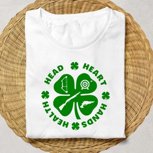 May include: White t-shirt with a green four-leaf clover design. The clover has a bow and arrow, a target, a rifle, and a pistol inside. The text around the clover reads "Head, Heart, Hands, Health".