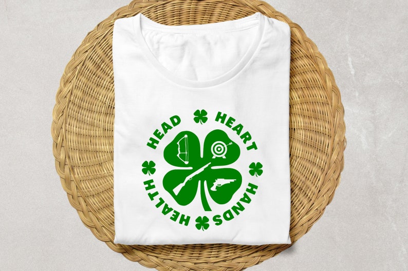 May include: White t-shirt with a green four-leaf clover design. The clover has a bow and arrow, a target, a rifle, and a pistol inside. The text around the clover reads "Head, Heart, Hands, Health".