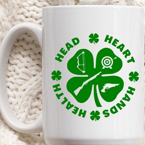 May include: White ceramic mug with a green four-leaf clover design. The clover has a bow and arrow, a target, a rifle, and a pistol in each leaf. The text around the clover reads "Health, Head, Heart, Hands".