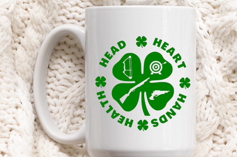 May include: White ceramic mug with a green four-leaf clover design. The clover has a bow and arrow, a target, a rifle, and a pistol in each leaf. The text around the clover reads "Health, Head, Heart, Hands".