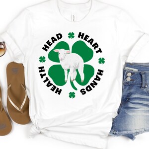 May include: White t-shirt with a green four-leaf clover design. The clover has a white lamb in the center. The text around the clover reads "Head, Heart, Hands, Health".