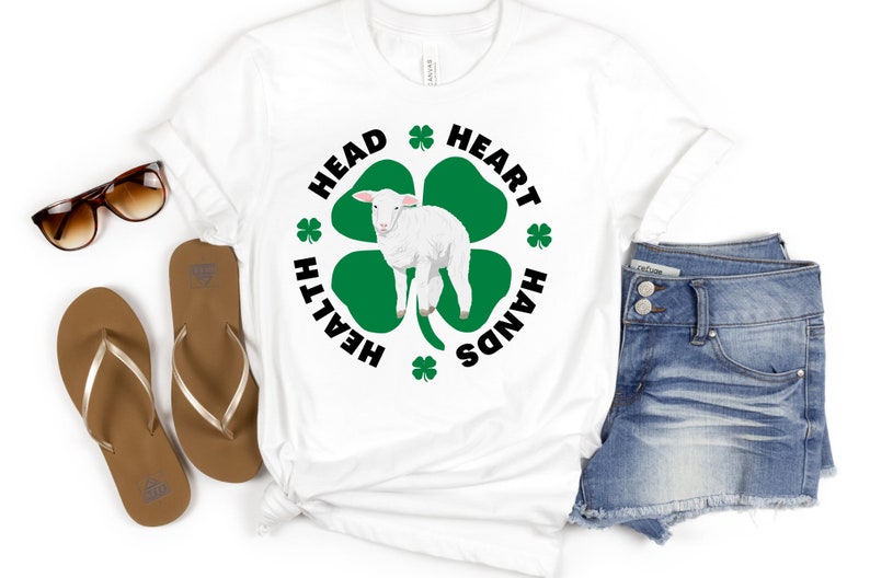 May include: White t-shirt with a green four-leaf clover design. The clover has a white lamb in the center. The text around the clover reads "Head, Heart, Hands, Health".