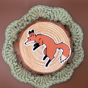 May include: A sticker of a cartoon fox in shades of orange, white, and black, leaping across a wooden slice. The wooden slice is set on a light green crocheted coaster. The background is a solid brown.